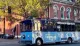 A blue trolley bus with advertising wraps for The New Franklin tour is seen on a sunny street with a backdrop of a brick building and a traffic signal showing red.