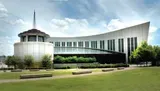 The image shows the Country Music Hall of Fame and Museum, with its modern architectural design and distinctive windows, located in a large open space with grass and trees under a partly cloudy sky.