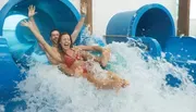 Two people are joyfully sliding down a water chute into a splash pool, with their arms raised and expressions filled with excitement.