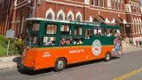 A green and orange trolley bus labeled Old Town Trolley Tours is carrying passengers on a hop-on hop-off tour in a city street.