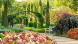 A serene garden pathway is flanked by lush greenery and vibrant flowers with a metal arch and a wooden bench inviting visitors to enjoy the surroundings.
