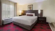 The image shows a neatly arranged hotel room with a large bed, a patterned carpet, and simple decor.