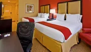 The image displays a bright and neatly arranged hotel room with two queen-sized beds, a work desk and chair, and warm color accents.