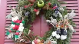 This image features an elaborate and festive Christmas wreath adorned with greenery, red berries, pine cones, a large green and red bow, and what appears to be a pair of miniature boots, displayed on a pastel burgundy door.