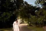 A person in a white outfit is walking down a dimly lit tree-lined path at dusk or night.