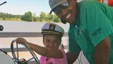 A man and a child, both smiling, are steering a boat with the child wearing a captain's hat.