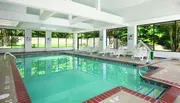 The image shows a serene indoor swimming pool with a row of lounging chairs by the side, natural light streaming in from ample windows, and reflections on the water surface suggesting tranquility.