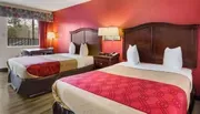 The image displays a neatly arranged hotel room with two queen-size beds, a bold red accent wall, and simple decor, suggesting a comfortable and straightforward accommodation.