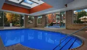 The image depicts an indoor swimming pool with a skylight and large windows offering a view of an outdoor patio area and building exterior.