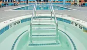A symmetrical view of stainless steel pool steps leading down into a shallow section of a pool with clear blue water, marked with depth indicators.
