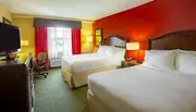 The image shows a bright and colorful hotel room with two beds, a red accent wall, and a view out the window.