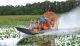 A group of people are joyfully waving while riding an airboat through a waterway with lush greenery on the sides.