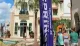 The image captures a sunny day at an outdoor shopping area with visitors walking past a banner, palm trees, and a small fountain.