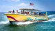 A group of people enjoys a sunny day on a speeding yellow boat named Sea Screamer on the water with an American flag flying at the stern.