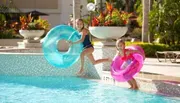 Two children are joyfully jumping into a swimming pool with inflatable tubes.