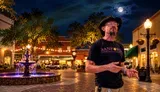 A man wearing a cowboy hat and a Sanford Tours & Experiences t-shirt is speaking while gesturing with his hands, standing in an outdoor plaza with a fountain and string lights in the blurred background.