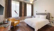The image shows a modern and stylish hotel room with brick walls, a large bed, chic furnishings, and plenty of natural light.