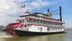 The image shows a classic multi-deck riverboat with a large red paddle wheel, named City of New Orleans, docked under a clear sky with American flags fluttering atop.