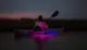 A person is kayaking in tranquil waters at dusk with a striking purple light illuminating the water beneath the kayak.