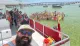 A group of people is joyfully posing for a selfie on a boat while another crowd in the water behind them is also partaking in the photo, all set against a backdrop of a sunny, busy waterway filled with boats.