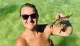 A smiling woman in sunglasses is standing in clear green water, holding a small crab up to the camera.