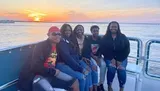 A group of five people are smiling and posing together on a boat with a beautiful sunset in the background.