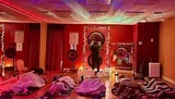 The image shows a dimly-lit room with a cosmic star projector effect on the ceiling, while several people lay on mats under blankets, and a person at the front appears to be performing a sound healing or meditation session with large gongs.