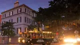 A Ghosts & Gravestones tour trolley is filled with passengers and travels through a street at dusk, with a historic building to the left and trees draped with Spanish moss overhead.