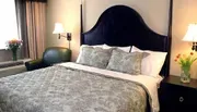 The image shows a neatly made bed with a floral patterned comforter, flanked by matching nightstands with lamps, against an elegant black headboard in a well-lit room with fresh flowers adding a pop of color.