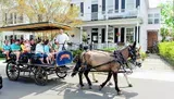 A horse-drawn carriage is carrying passengers on a sunny day past residential buildings, guided by a coachman.
