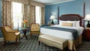 The image shows an elegantly furnished hotel room with a queen-sized bed, blue walls, patterned curtains, and classic-styled furniture.