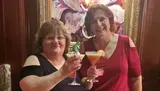 Two smiling women are toasting with cocktails in front of a vibrant abstract painting.