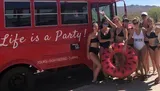 A group of people in swimwear is posing cheerfully beside a red bus with the slogan Life is a Party! and the iconic Route 66 sign, indicating a fun, social outing probably linked to a bachelorette celebration as indicated by one person wearing a Bride swimsuit.