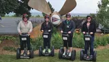 Four people wearing helmets are standing in a line on Segways in front of a large airplane propeller sculpture.