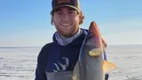 A man in a cap and hoodie is smiling while holding a large fish in front of a snowy or icy landscape.