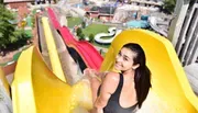 A person is sitting at the top of a water slide, smiling back at the camera with a water park in the background.