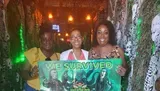 Three people are smiling and holding a sign stating WE SURVIVED, with a horror-themed backdrop that includes eerie green lighting and skulls, suggesting they completed a scary entertainment attraction.