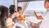A family is enjoying a scenic view from inside a Ferris wheel gondola, with a sunny coastal cityscape stretching out in the background.