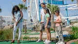 A family of four is enjoying a game of mini-golf on a sunny day with a ferris wheel and other attractions in the background.