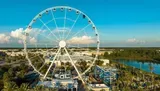 The image shows a large observation wheel towering over a scenic area with clear blue skies, lush greenery, and a blue waterbody nearby.