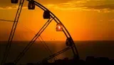 The image features the silhouette of a Ferris wheel against a striking orange sunset sky over the ocean horizon.