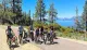A group of people are posing with their bicycles on a sunny day near a picturesque lake surrounded by mountains and trees.