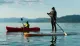 Two people are enjoying water sports on a calm lake with mountains in the background, one kayaking and the other paddleboarding.