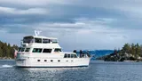 A white pleasure boat with people on deck is cruising on the water with a backdrop of pine trees and a cloudy sky.