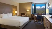 The image shows a neatly arranged hotel room with modern furnishings and a large window offering a stunning view of snow-capped mountains and a pine forest.