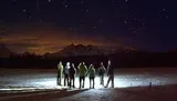 A group of people stand on a snow-covered ground at night, gazing at a starry sky with a backdrop of mountains.