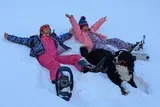 Two children and a dog are enjoying playing in the snow, with one child lying down and another sitting, both are wearing winter gear, while the dog stands beside them.