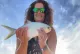A smiling woman wearing sunglasses holds up a fish to the camera against a bright sky backdrop.