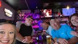 A group of people poses for a happy selfie at a bar with television screens and an array of liquor bottles in the background.