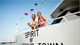 A man and a woman are standing beside the railing of a boat named SPIRIT OF HARROW TOWN, adorned with colorful flags, looking out towards the horizon.